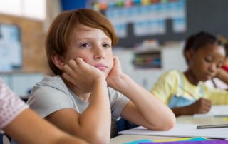 Tired School Boy With Hand on Face Sitting at Desk in Classroom